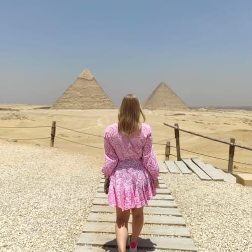 A woman in a pink dress walking on a wooden pathway towards the Pyramids of Giza in Egypt.