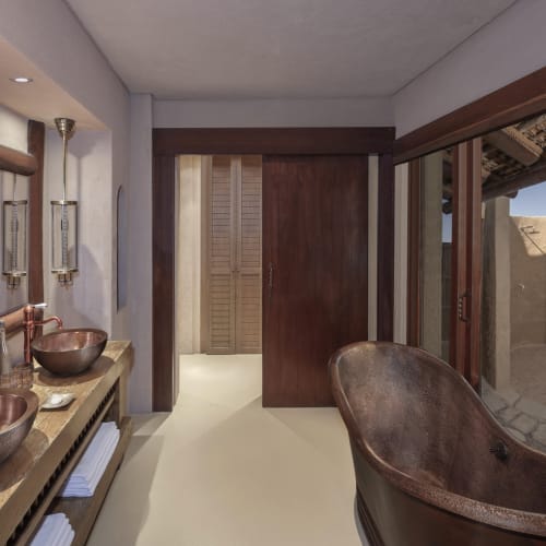Luxuriöses Badezimmer mit Kupferwaschbecken, großer freistehender Kupferbadewanne und Holzakzenten, mit offenem Fensterblick auf einen Außenbereich mit rustikalen Wänden.