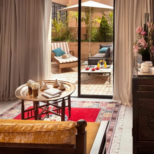 Ein gemütlicher Wohnbereich in einer Suite mit einem Holzcouch mit gelben Kissen, einem runden Holztisch mit Dekorationsgegenständen und einer offenen Glastür, die zu einer Terrasse mit Liegestühlen und einem weißen Sonnenschirm führt.