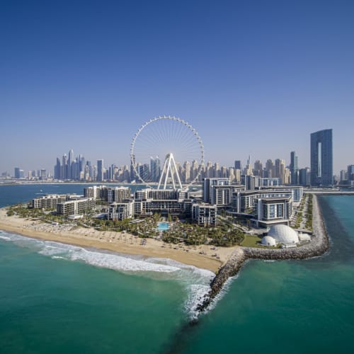 Aerial view of Dubai's coastline featuring a large Ferris wheel, modern buildings, and a sandy beach with the city skyline in the background.
