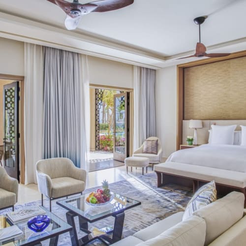 Spacious hotel room with a large bed, seating area, and balcony doors leading to an outdoor seating area with greenery.