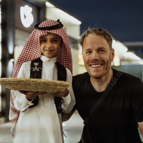 A young boy in traditional Middle Eastern attire holding a tray, standing next to a smiling man in casual clothing at an airport.