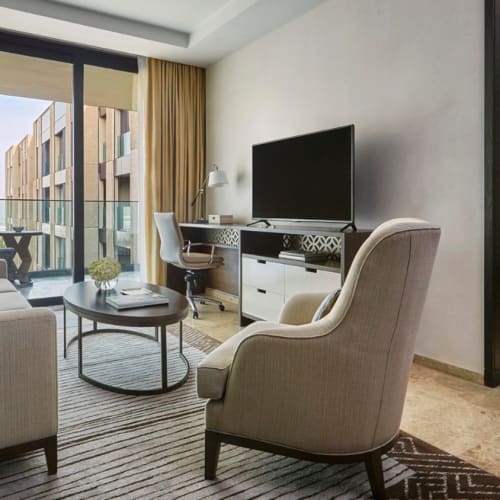Modern hotel living room with a balcony view, featuring a sofa, armchair, coffee table, TV, and a bedroom visible through an open doorway.