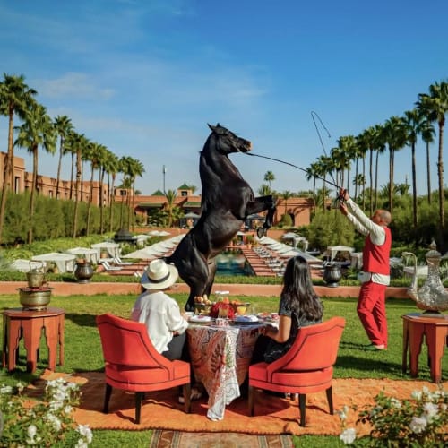 Menschen genießen ein Frühstück im Freien in einem Garten mit einer aufspringenden Pferdestatue und Palmen im Hintergrund.