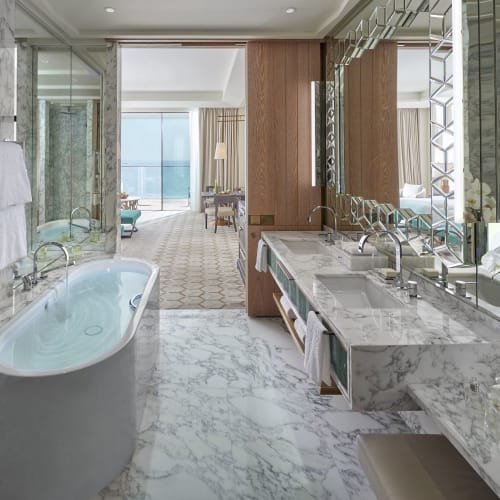 Luxushafte Hotelsbad mit Marmorflächen, Badewanne und Doppelwaschbecken, das zu einem hellen Schlafzimmer und Balkon führt.