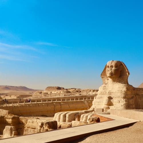 Die große Sphinx von Gizeh steht deutlich sichtbar vor klarem blauen Himmel, mit den Pyramiden im Hintergrund bei hellem Sonnenlicht.