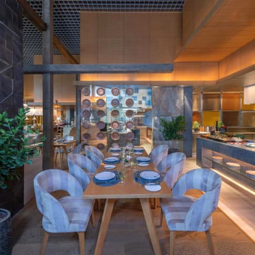 Elegantes Restaurantinnere mit einem hölzernen Esstisch für acht Personen, Samt-Stühlen, Topfpflanze und einem offenen Küchenbereich im Hintergrund.