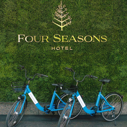 Two blue rental bicycles parked in front of a lush green wall with the Four Seasons Hotel logo in gold lettering above them.