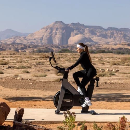 A woman is riding an exercise bike outdoors on a paved trail in a desert landscape with mountains in the background.