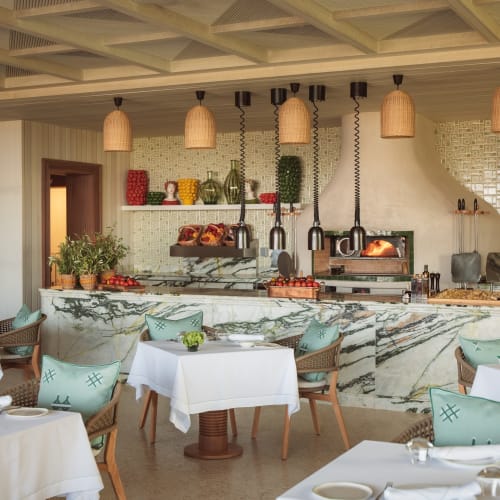Elegantes Restaurantinnenraum mit Tischen mit weißen Tischdecken, hell-türkisfarbene Stühle und einer Marmortheke mit Grill und hängenden Lampen.