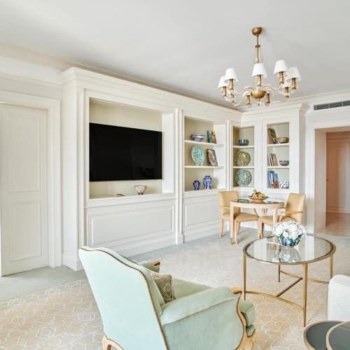 Luxurious hotel suite with elegant white furniture, a chandelier, and a television in the living area, with a bedroom visible through an open doorway.