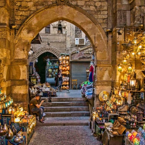 Ein lebendiger Marktplatz in Kairo mit Ständen, die bunte Lampen, Keramik und Metallwaren unter einem alten Steingang verkaufen.
