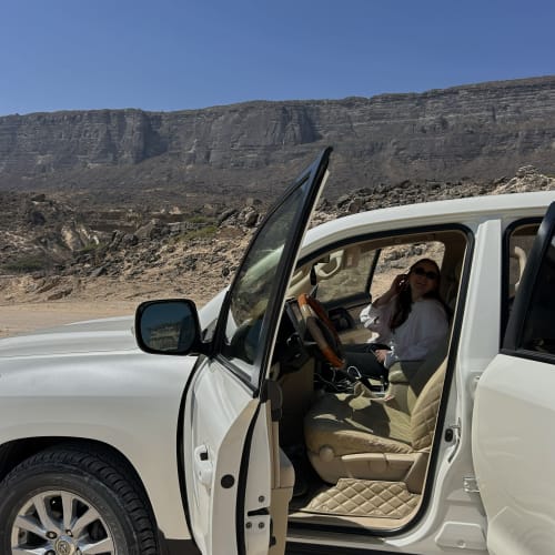 A woman sitting in the driver's seat of a white SUV with the door open, parked in a rocky desert landscape with steep cliffs in the background under a clear blue sky.
