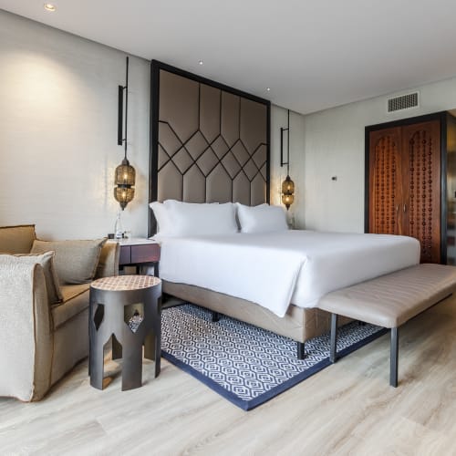 Modern hotel room with a large double bed, beige sofa, patterned rug, and wooden flooring, featuring Moroccan-style hanging lamps and a wooden partition separating the bedroom from the bathroom area.