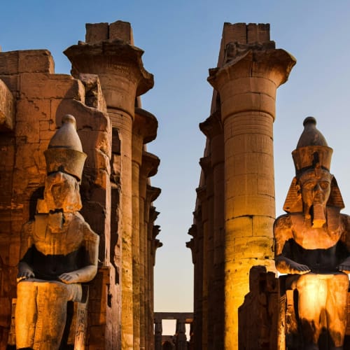 Alte ägyptische Tempelruinen mit großen Statuen und Säulen, hell erleuchtet bei Sonnenuntergang.