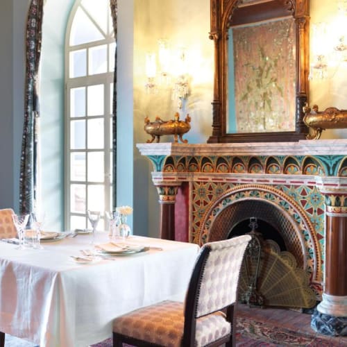 Ein Esstisch mit weißer Tischdecke, gedeckt mit Tellern und Gläsern, vor einem bunten, kunstvoll verzierten Kamin mit großem Spiegel darüber in einem eleganten Raum.
