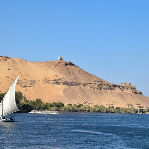 A sailboat gliding on the river in front of a hill with ancient ruins and a small structure at the top.