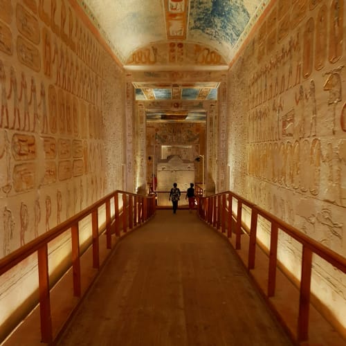 A corridor inside an ancient Egyptian tomb with walls and ceiling adorned with colorful hieroglyphs and wall carvings, with visitors walking towards the tomb chamber.