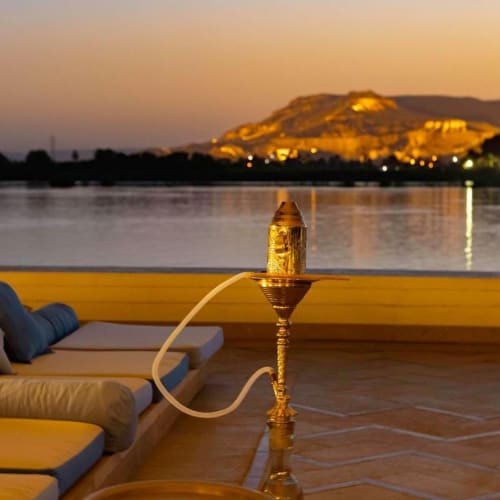 Luxurious lounge chairs and cushions overlooking the Nile River at sunset, with a traditional hookah in the foreground and a historical hill illuminated in the background.