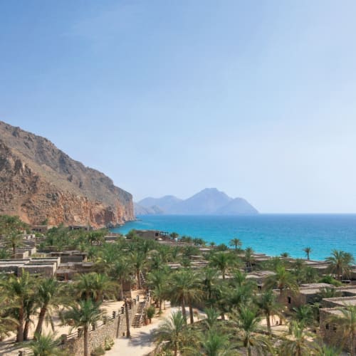 Eine Luftaufnahme des Resorts Six Senses Zighy Bay mit üppigem Grün, Palmen, bergigem Hintergrund und dem Meer im Hintergrund.