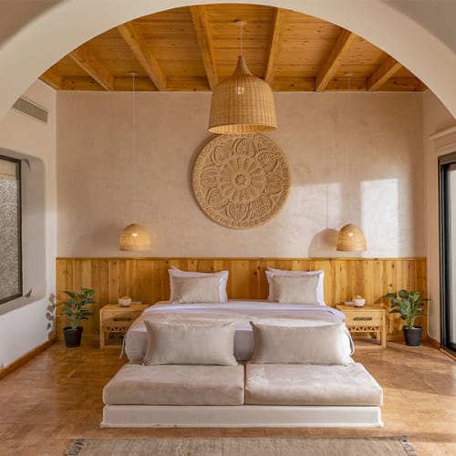 Cozy hotel suite bedroom with wooden ceiling, twin beds with white linens, wicker pendant lamps, decorative round wall art, and potted plants, viewed through an arched doorway.