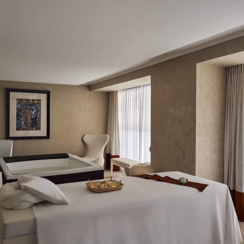 Ein geräumiger Hotel-Behandlungsraum mit einem Bett, das mit weißen Laken bedeckt ist, großen Fenstern mit transparenten Vorhängen und eleganter Dekoration.