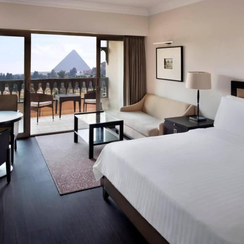 Ein Hotelzimmer mit Balkon mit Blick auf die Pyramiden von Gizeh in Kairo, mit großem Bett, Sofa und modernen Möbeln.
