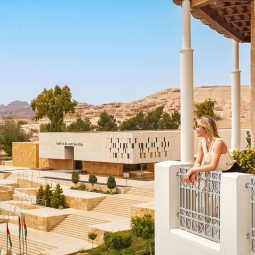 Eine Frau sitzt auf einem Balkon mit Blick auf ein modernes Gebäude mit arabischer Schrift und Wüstenlandschaft in Petra, Jordanien.