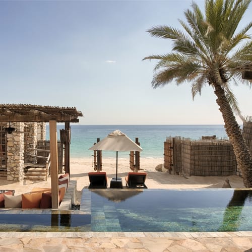 Ein luxuriöser Poolblick mit Blick auf einen Strand, eine Palme, schattige Sitzgelegenheiten und den Ozean im Hintergrund.