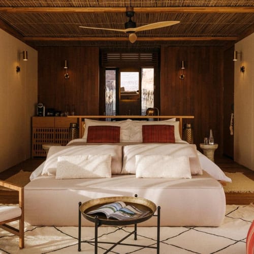 Ein gemütliches Schlafzimmer mit großem Bett, Holzpaneele an der Wand, weicher Beleuchtung, Deckenventilator und ein Fenster, das Tageslicht einlässt.