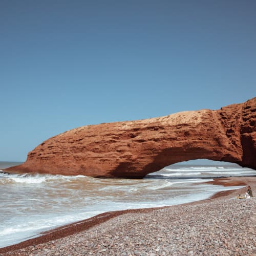 A natural rock formation on a beach with a large archway opening, under a clear blue sky.