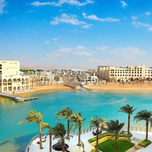 Luxurious hotel complex with a large water body, palm trees, and modern architecture under a bright blue sky.