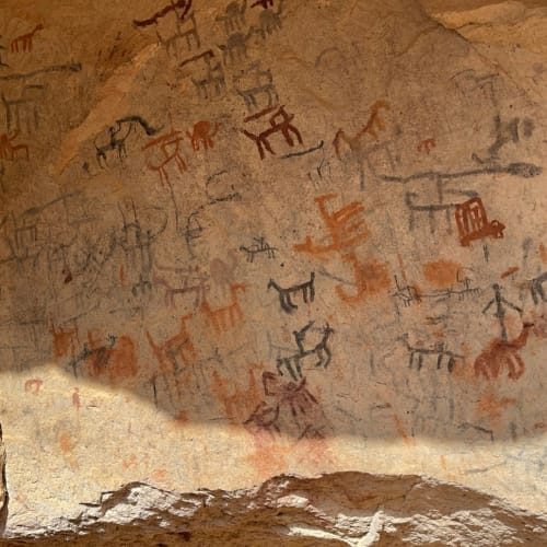 Alte Höhlenwand mit bunten Felszeichnungen und Inschriften, teilweise im natürlichen Licht beleuchtet.