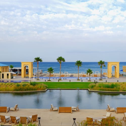 Luxuriöses Resort mit gelben Gebäuden, Palmen, Pools und einem Sitzbereich am Wasser an einem ruhigen Gewässer mit Blick auf das Meer unter leicht bewölktem Himmel.