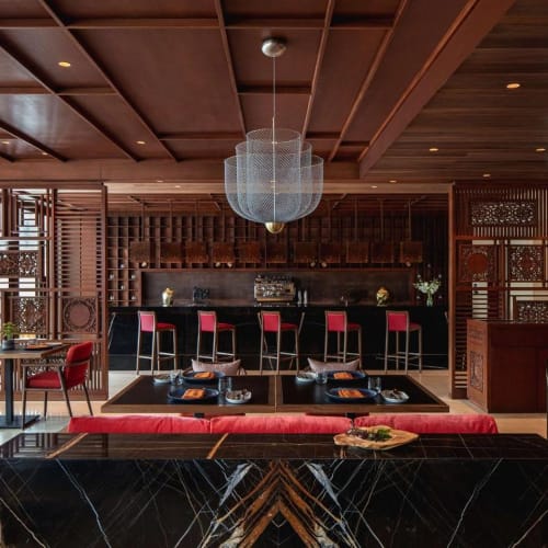 Elegant restaurant with a modern interior featuring a bar with pink stools, black tables, and red chairs, illuminated by a large central chandelier.