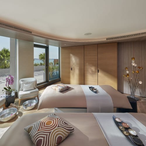 Luxuriöses Hotelzimmer mit zwei ordentlich gemachten Betten, Sitzecke mit Stühlen und großen Fenstern mit Blick auf Palmen und die Stadt.