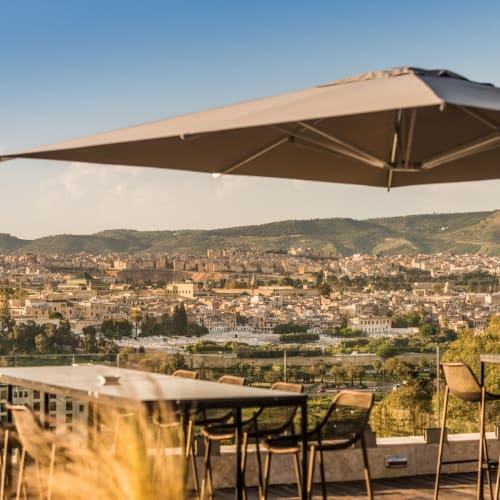 Außenterrasse einer Hotelbar mit Tischen, Stühlen und großem Sonnenschirm mit Blick auf eine Stadtlandschaft umgeben von Hügeln unter klarem Himmel.