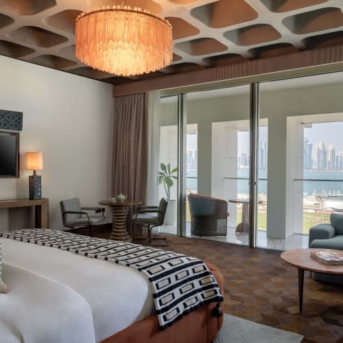 Geräumiges Hotelzimmer mit großem Bett, gemustertem Überwurf, Sitzecke und bodentiefen Fenstern mit klarem Blick auf das Meer und die Stadt Skyline am anderen Ufer.