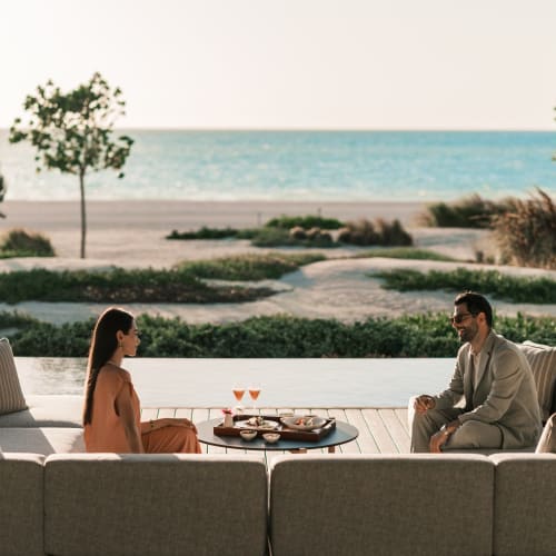 Ein Mann und eine Frau sitzen auf einem runden Sofa mit Blick auf den Strand und das Meer im Hintergrund.