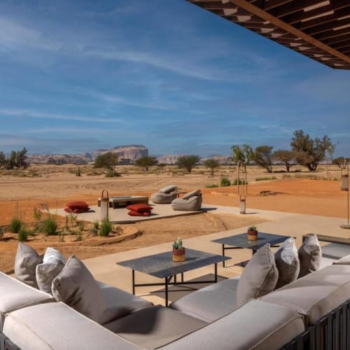 A luxurious outdoor seating area with a sofa and coffee tables on a terrace overlooking a desert landscape with sparse trees and rock formations under a blue sky.