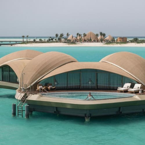 Luxuriöse Overwater-Villa mit geschwungenem Dach, Pool und Liegestühlen, auf türkisfarbenem Wasser gelegen, mit weiter Sicht auf andere Überwasserstrukturen und einen Sandstrand im Hintergrund.