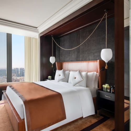 Luxury hotel bedroom with large bed dressed in white and brown linens, two white hanging lamps, floor-to-ceiling windows with city view, and a modern standing lamp.