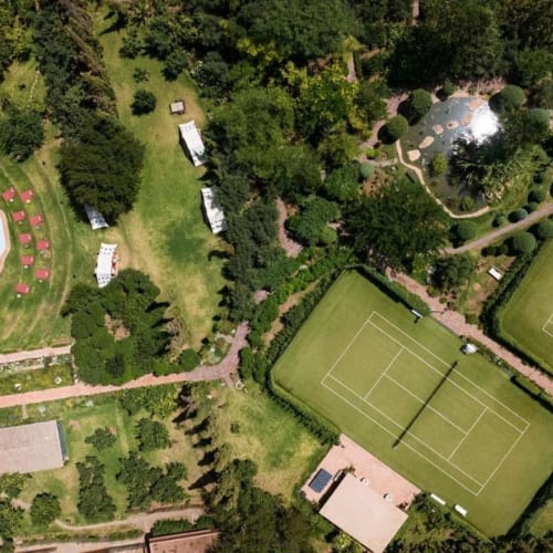 Luftaufnahme eines üppigen Gartens mit einem Schwimmbecken, Tennisplätzen und Wegen.