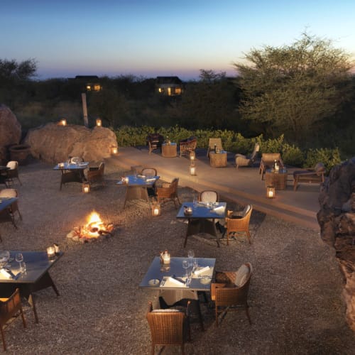 Außenbereich mit mehreren Tischen und Stühlen um eine zentrale Feuerstelle bei Dämmerung, umgeben von großen Felsen und Bäumen in einer natürlichen, wüstenähnlichen Umgebung.