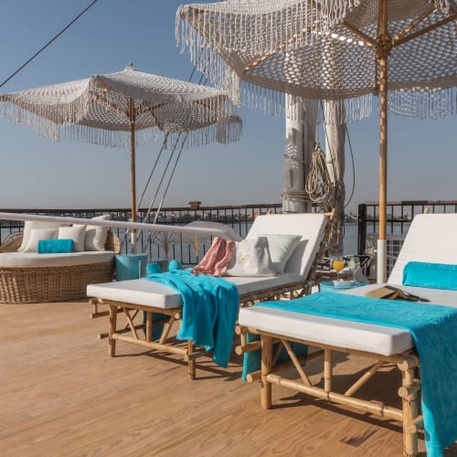 Two white cushioned sun loungers with blue towels and pillows on a boat deck, shaded by large white fringed parasols under a clear sky.