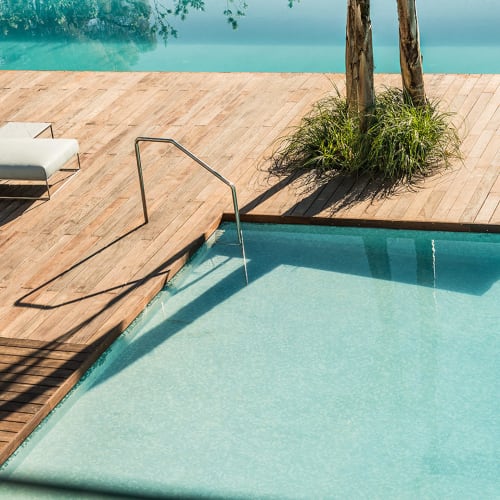 Ein moderner Swimmingpool mit klarem blauem Wasser und einem Holzdeck mit weißen gepolsterten Liegen und zwei von grünen Pflanzen umgebenen Bäumen.