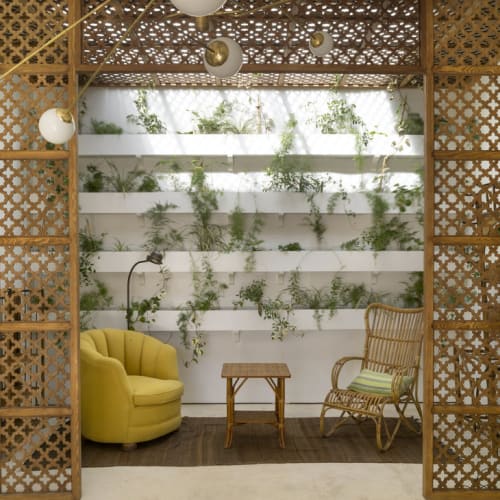 Gemütliche Außenterrasse mit einem gelben Sofa, Korbstuhl, kleinem Holztisch und einer vertikalen Gartenwand im Hintergrund, rahmenförmig durch dekorative Gitterpaneele und hängende Kugelleuchten.