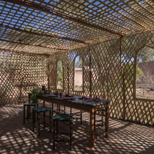 A rustic interior with a wooden table set for breakfast inside a lattice-woven structure, sunlight casting intricate shadows.