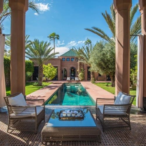 A luxurious private villa with a pool seen through classical arches, surrounded by lush greenery and palm trees.