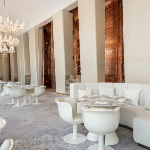 Heller und eleganter Speisebereich in der Masala Library des Fairmont Doha mit weißen, gepolsterten, geschwungenen Sofas, modernen weißen Stühlen, runden Tischen, Kristalllüstern und dekorativen Kupferwänden.